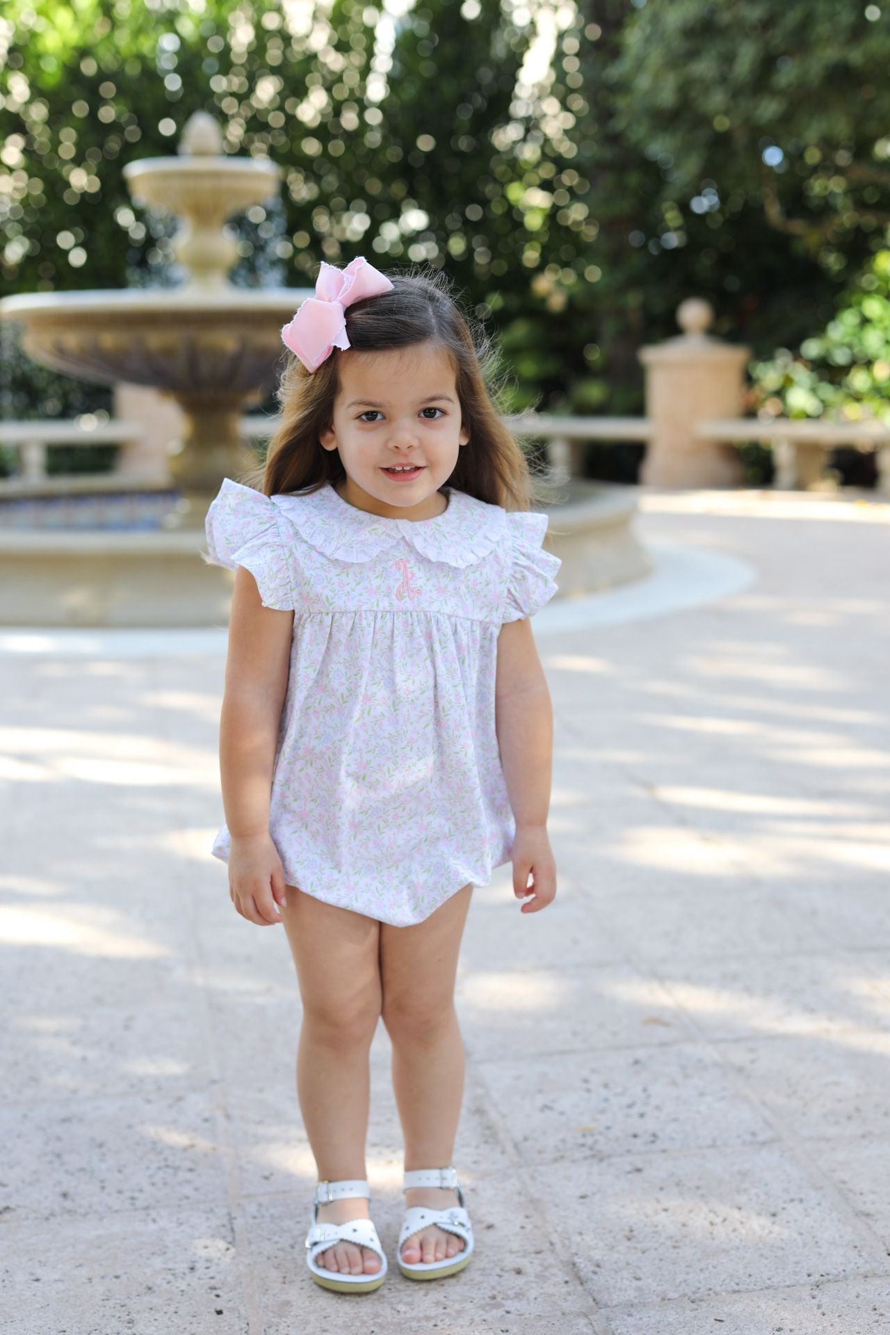 Young girl in a white dress standing in a sunlit outdoor setting with greenery.