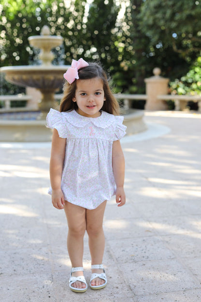 Young girl in a white dress standing in a sunlit outdoor setting with greenery.