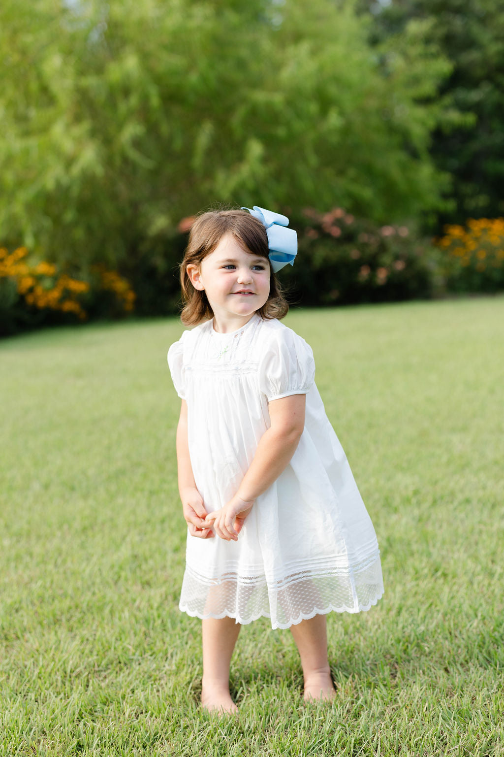 Young girl in a white dress standing on grass with greenery in the background