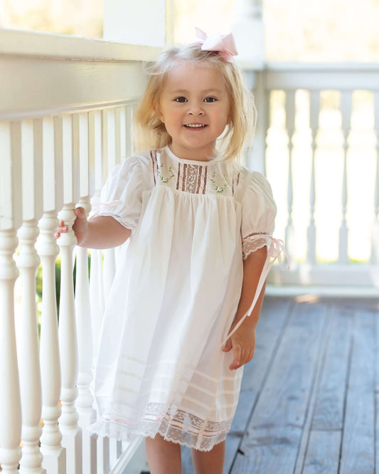 Young girl in a white dress with lace details standing on a wooden floor.