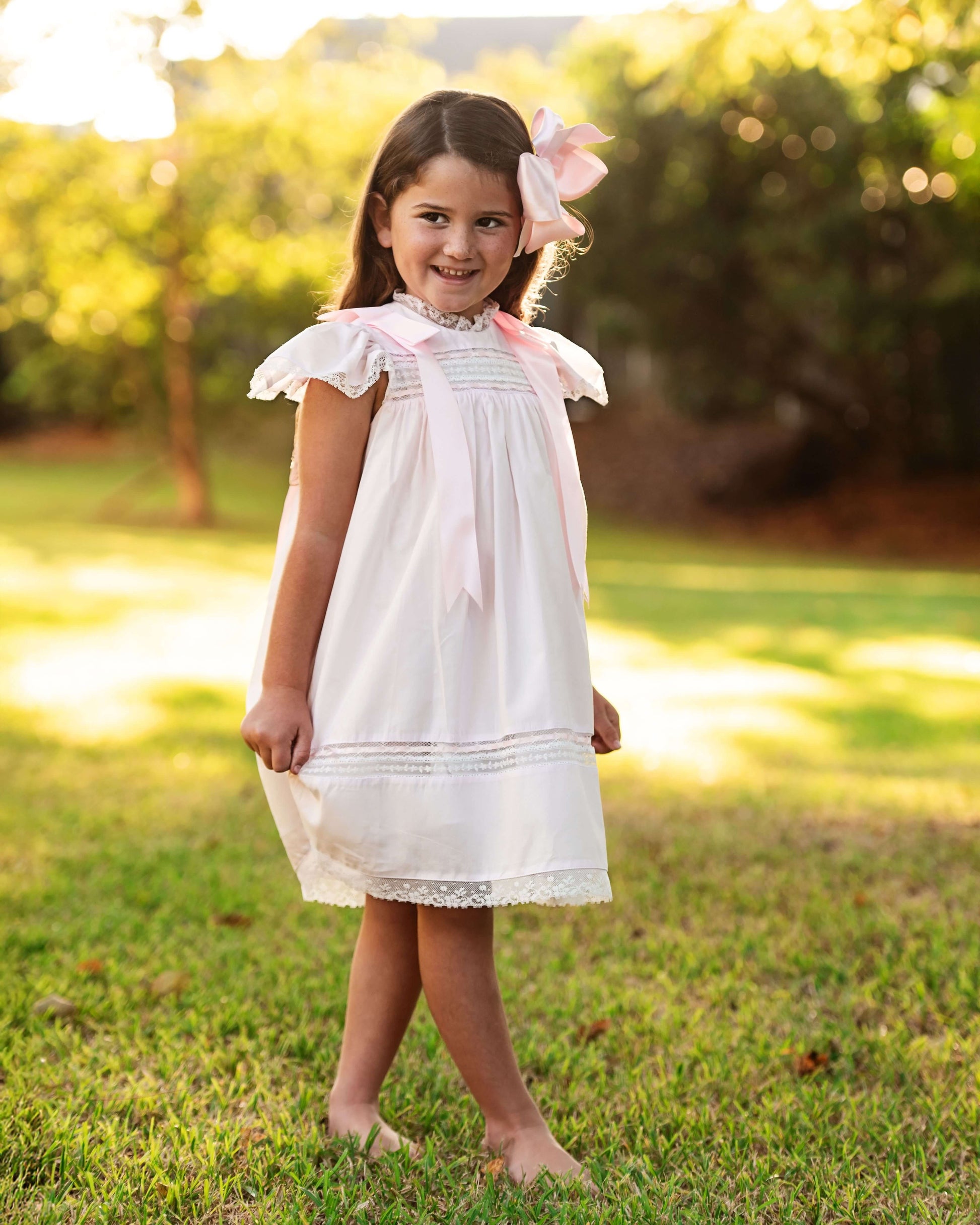Young girl in a white dress with pink accents standing in a grassy outdoor setting.