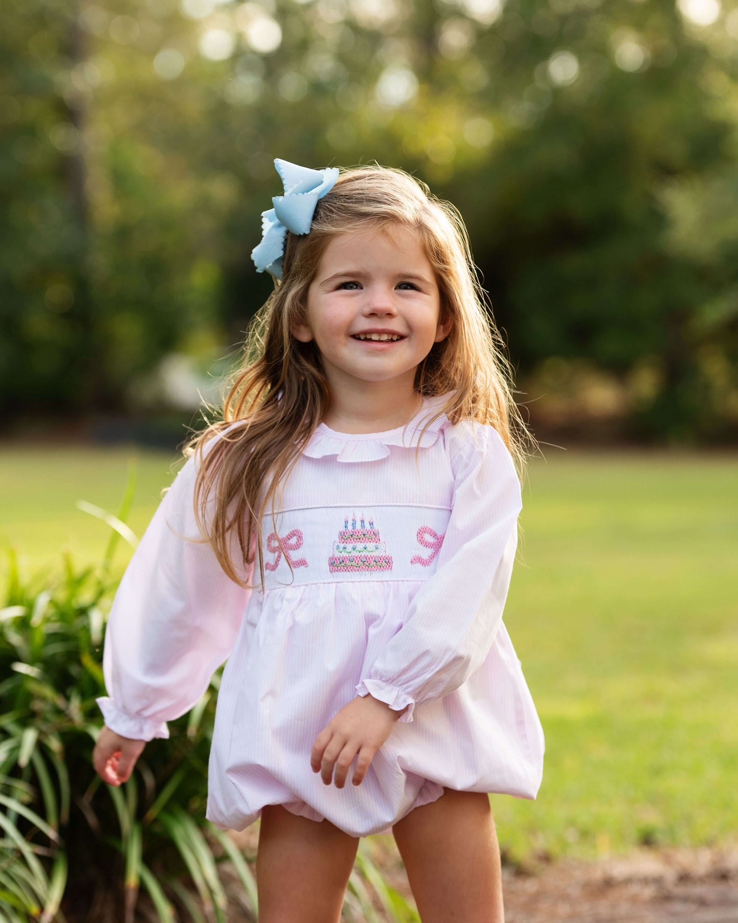 Young girl in a white dress with pink details standing outdoors in a natural setting.
