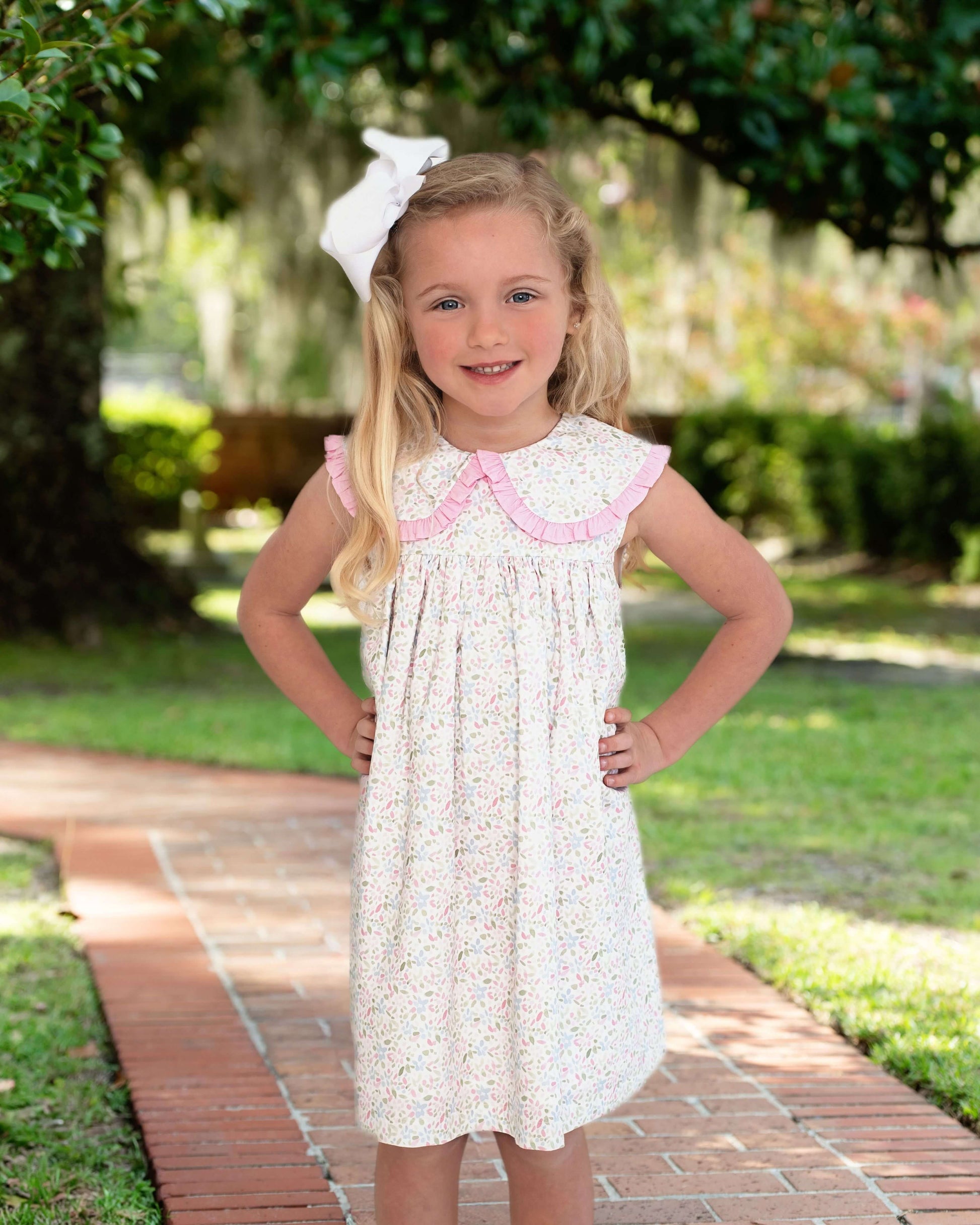 Young girl in a white dress with pink trim standing on a brick path in a garden.
