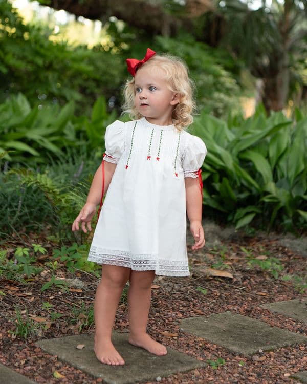 Young girl in a white dress with red accents standing on a stone path surrounded by greenery.