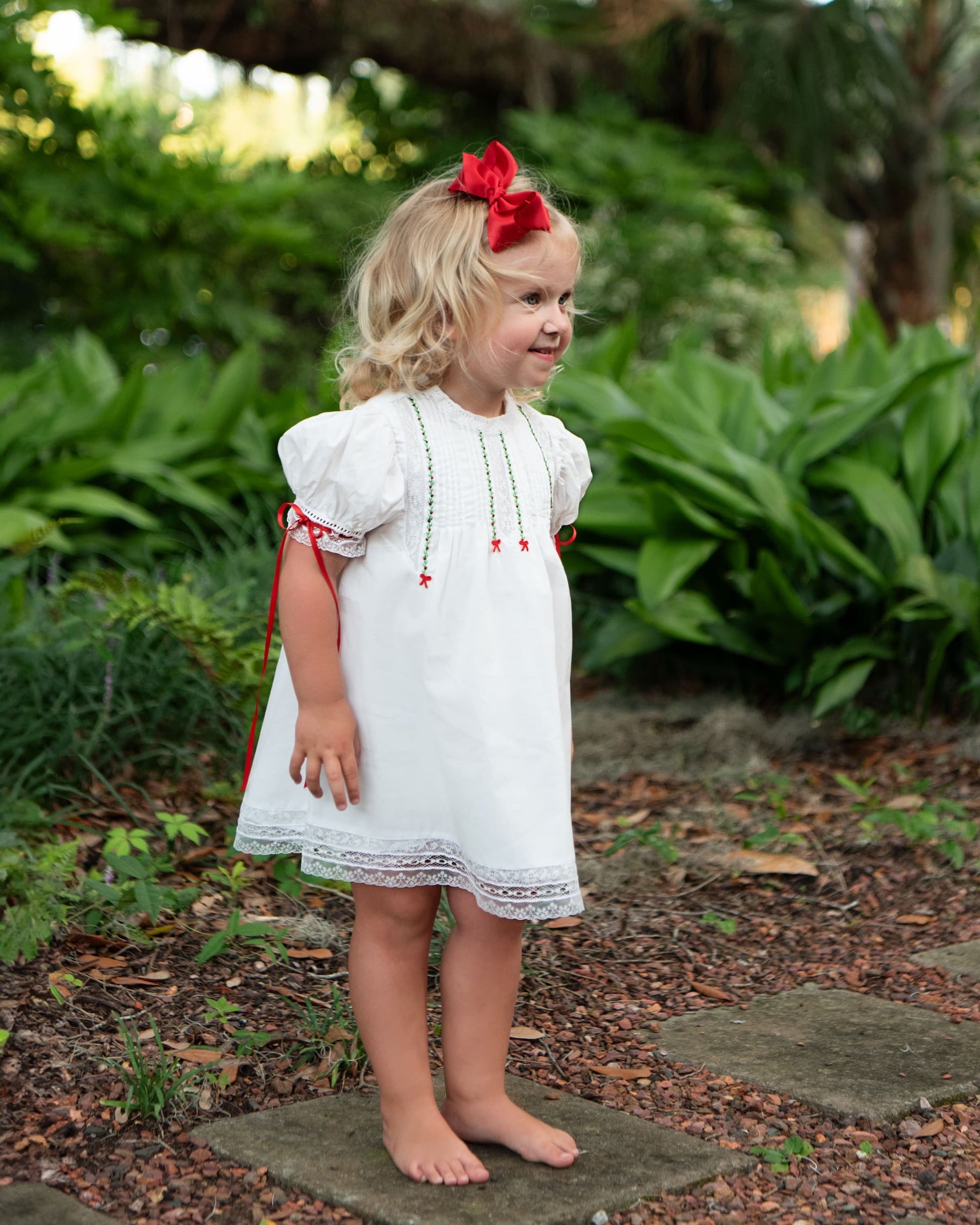 Young girl in a white dress with red accents standing outdoors among greenery.