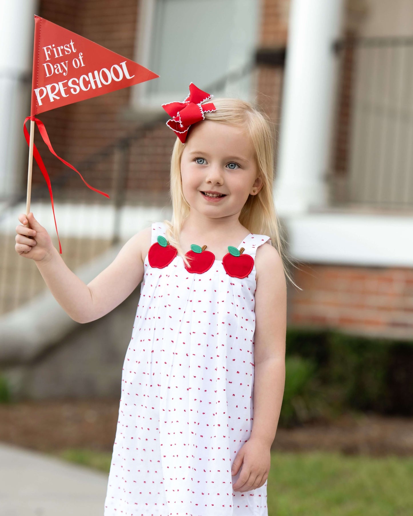 Young girl in a white dress with red apple patterns, holding a 'First Day of Preschool' flag.