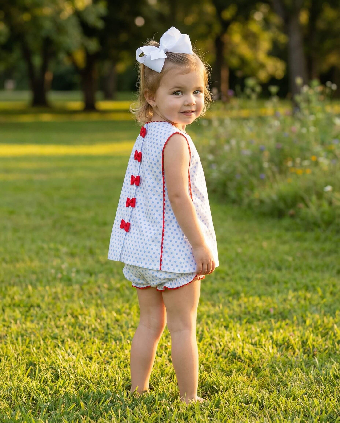 Young girl in a white dress with red buttons standing in a grassy field.