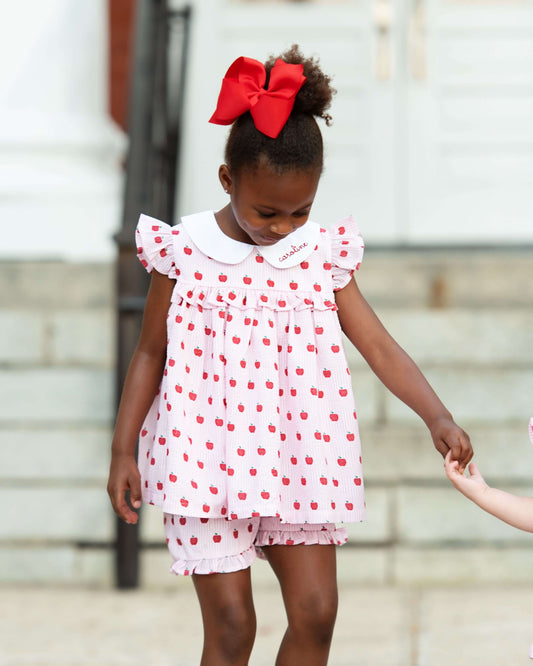 Young girl in a white dress with red polka dots and a large red bow standing outdoors.