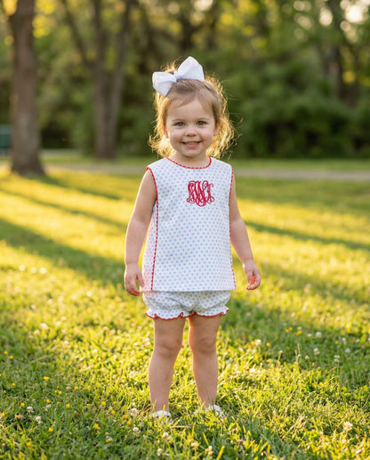 Young girl in a white dress with red trim and monogram standing in a grassy field.
