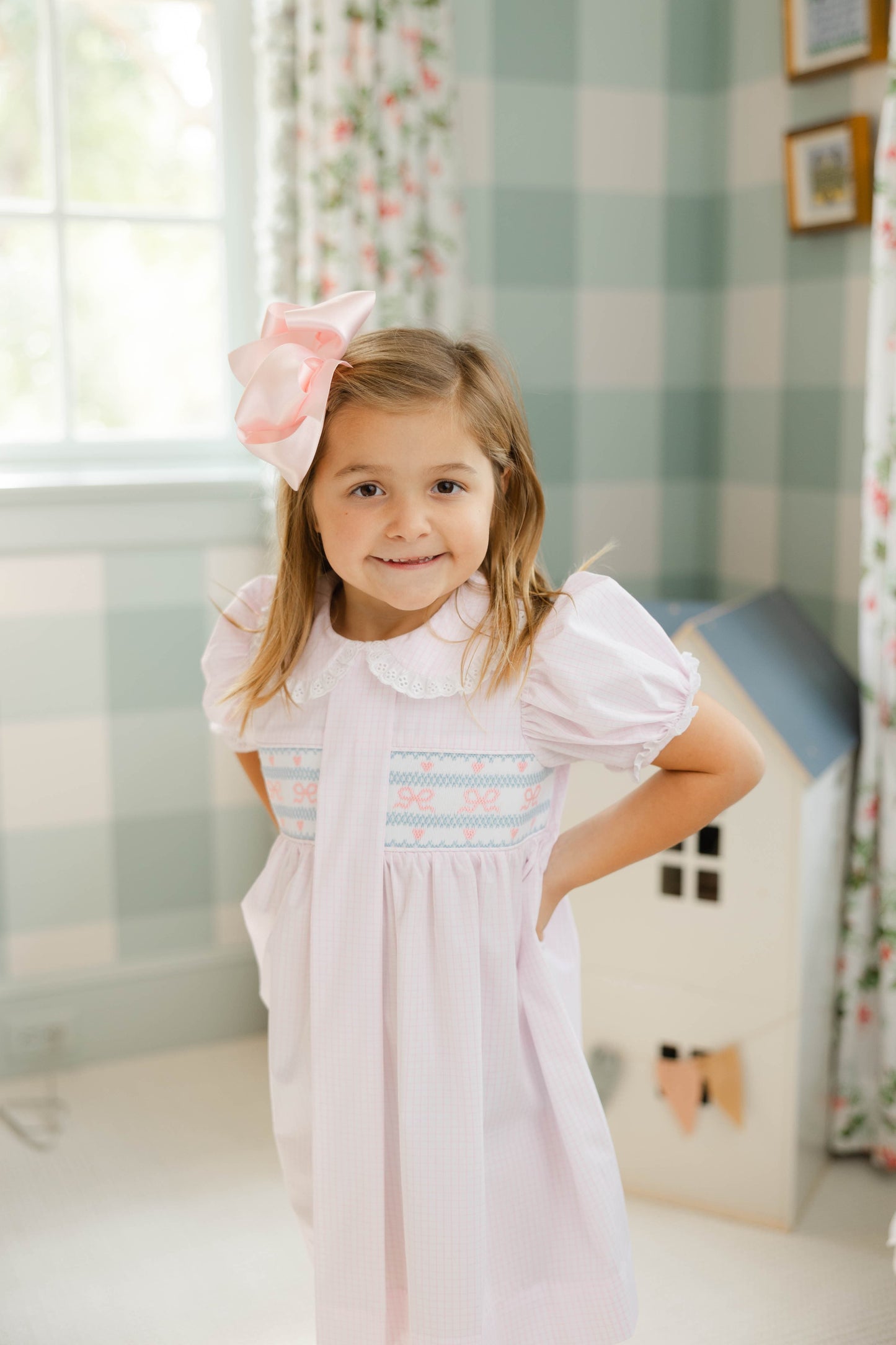 Young girl in a white dress with ruffled collar and sleeves, standing in a room with floral curtains and a checkered wall.