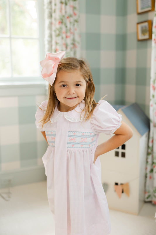 Young girl in a white dress with ruffled collar and sleeves, standing in a room with floral curtains and a checkered wall.