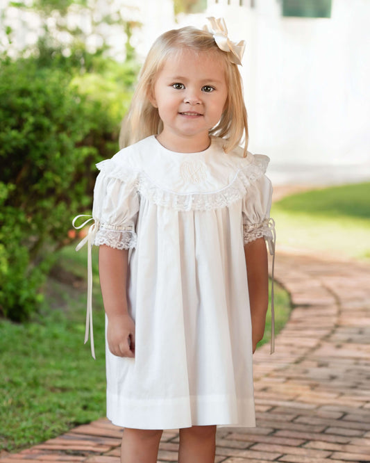 Young girl in a white dress with ruffled sleeves standing on a brick path with greenery in the background.