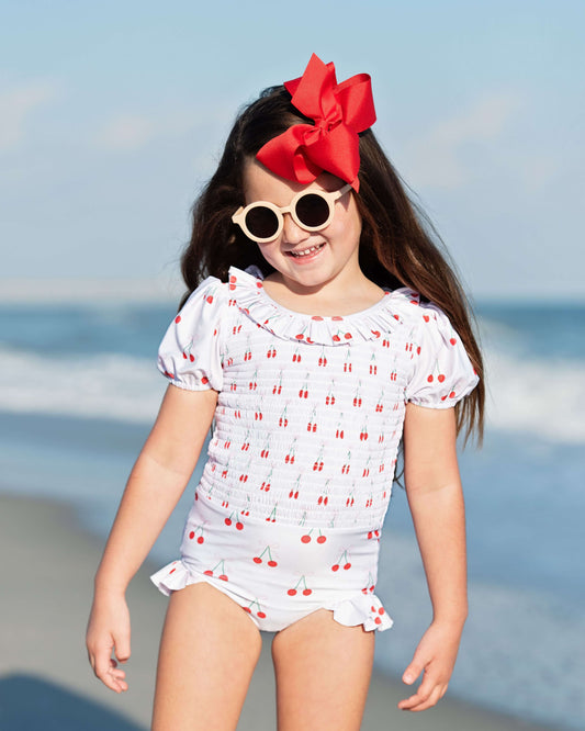 Young girl in a white swimsuit with red polka dots and a large red bow on her head, standing on a beach.