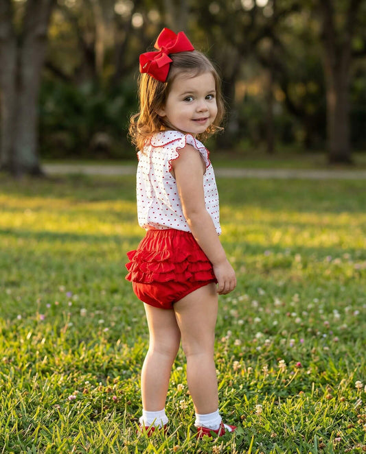 Young girl in a white top and red skirt with a red bow standing in a grassy field.