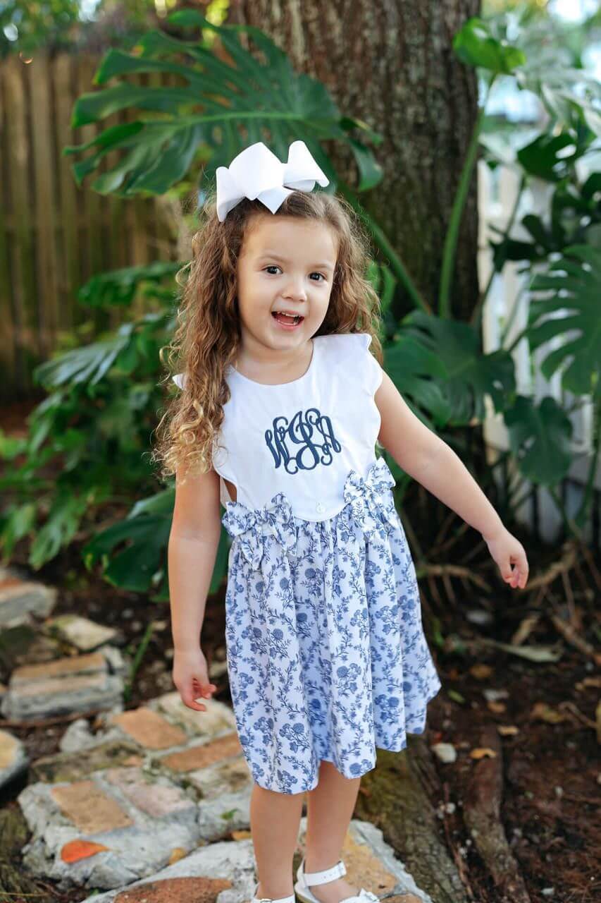 Young girl in a white top with monogram and blue floral dress standing outdoors among greenery.