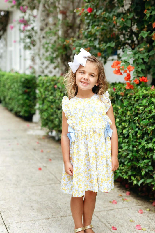 Young girl in a yellow floral dress with blue bows standing outdoors near greenery and flowers.