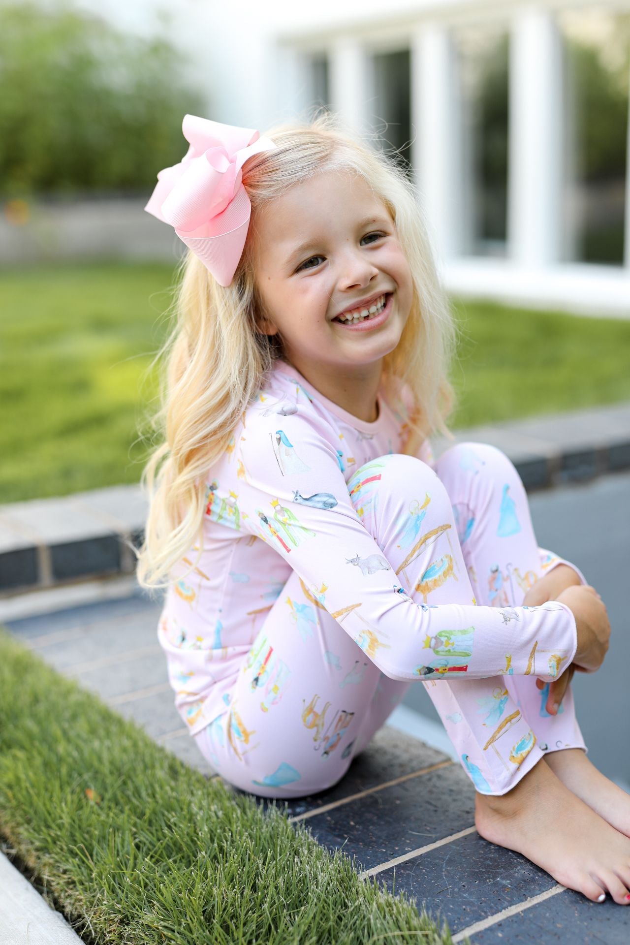 Young girl in pink pajamas with a large bow sitting on a stone path outdoors.