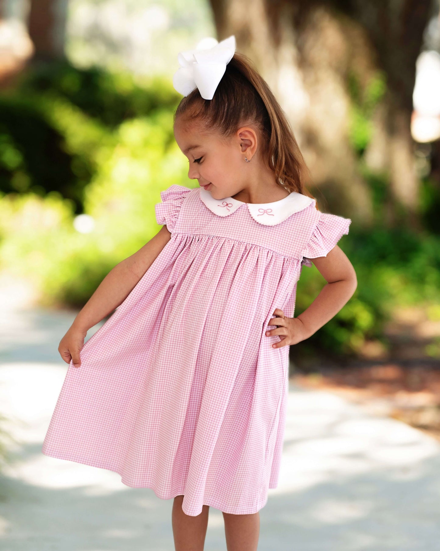 Young girl posing in a pink gingham dress with embroidered bows on the collar, standing outside