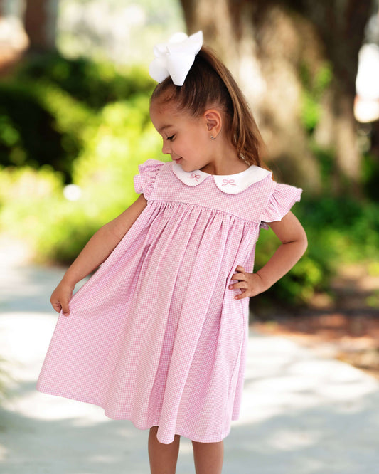 Young girl posing in a pink gingham dress with embroidered bows on the collar, standing outside