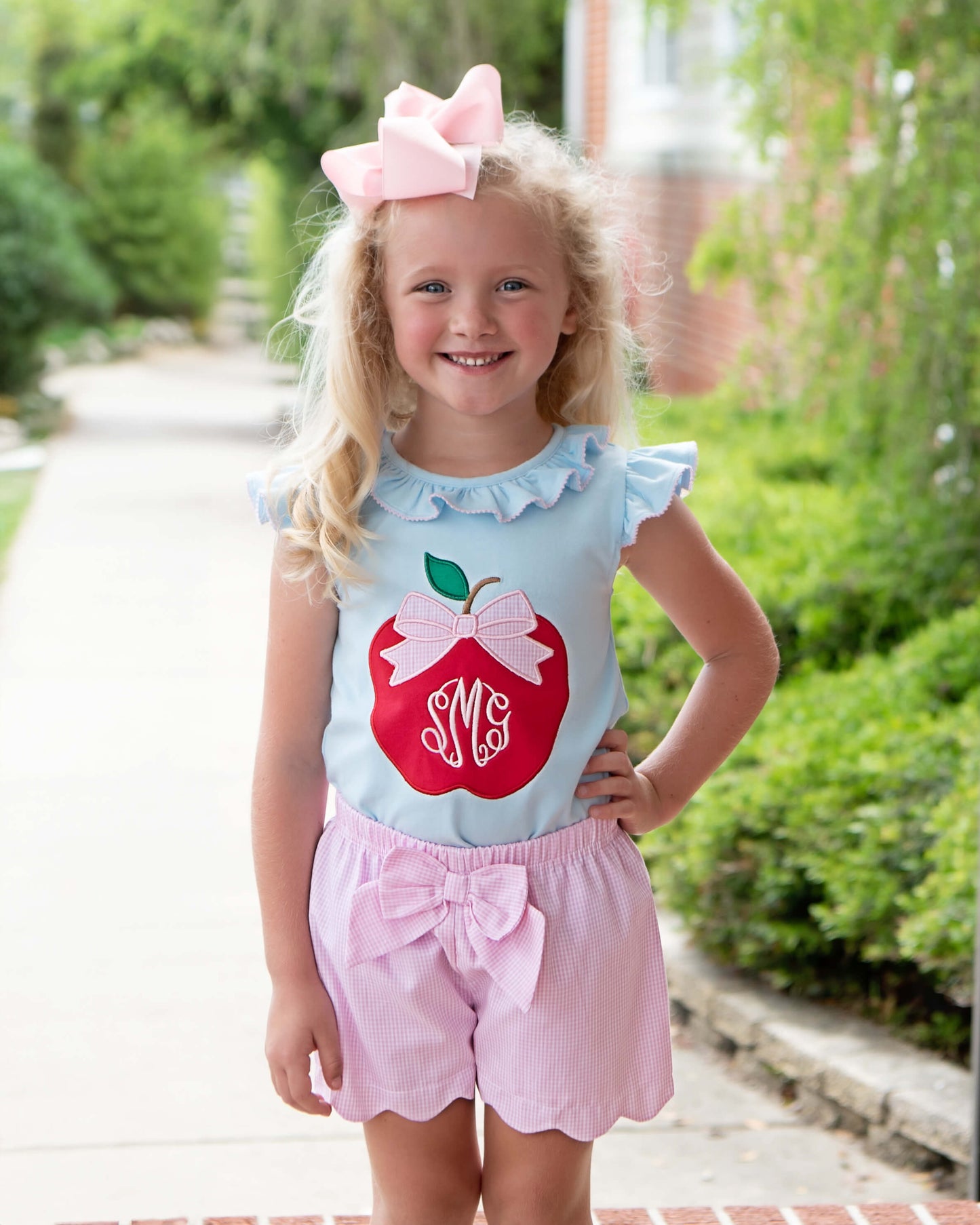 Young girl posing with hand on her hip wearing pink shorts and a shirt with an apple on it. 