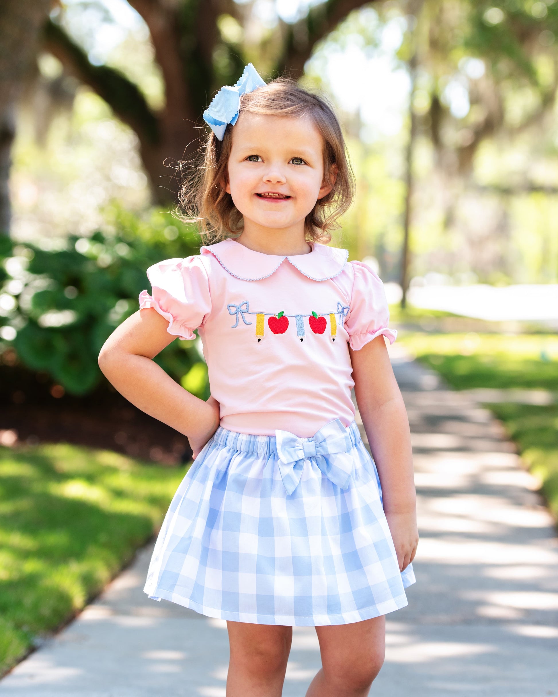 Young girl posing with her hand on her hip wearing a blue check skirt and pink shirt with pencils and apples.