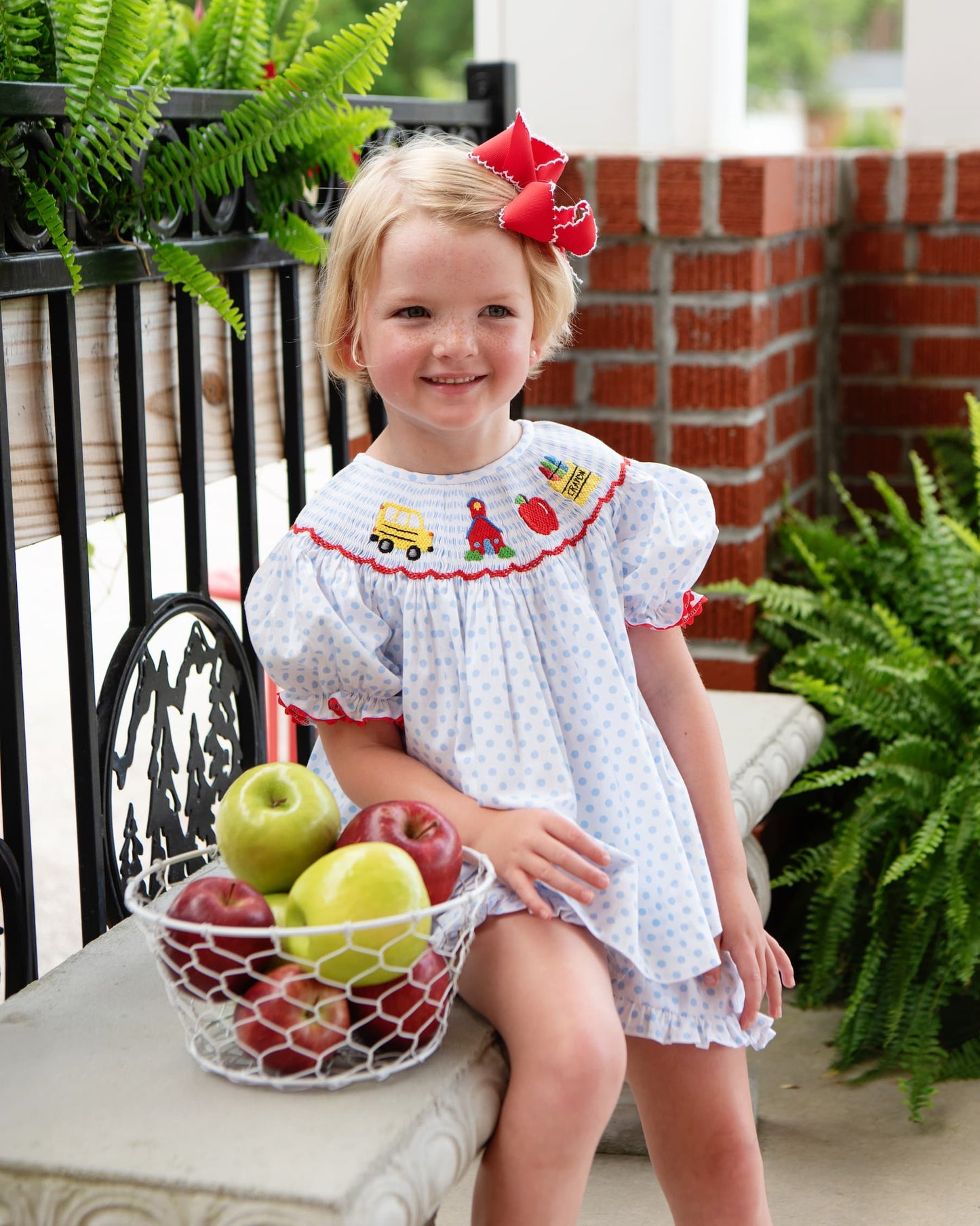 Young girl sitting on a bench next to a basket of apples wearing a blue dotted back to school outfit.