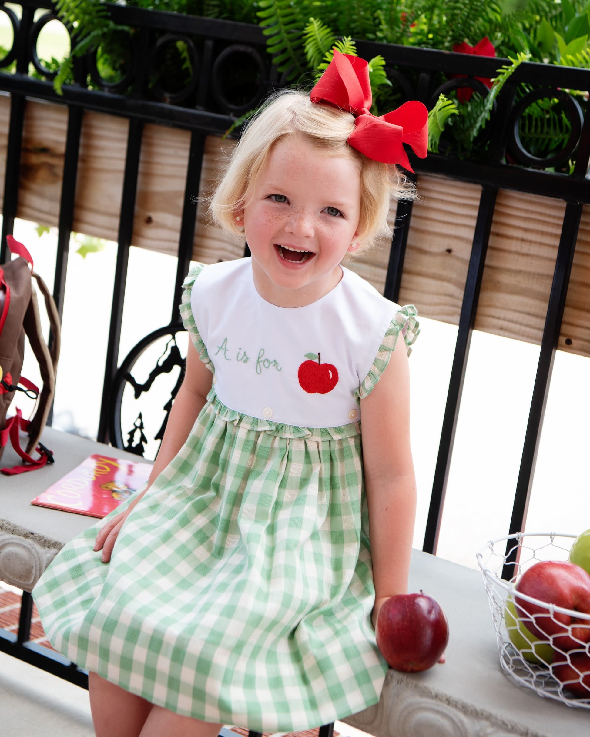 Young girl sitting on a bench next to apples wearing an A is for apple dress.