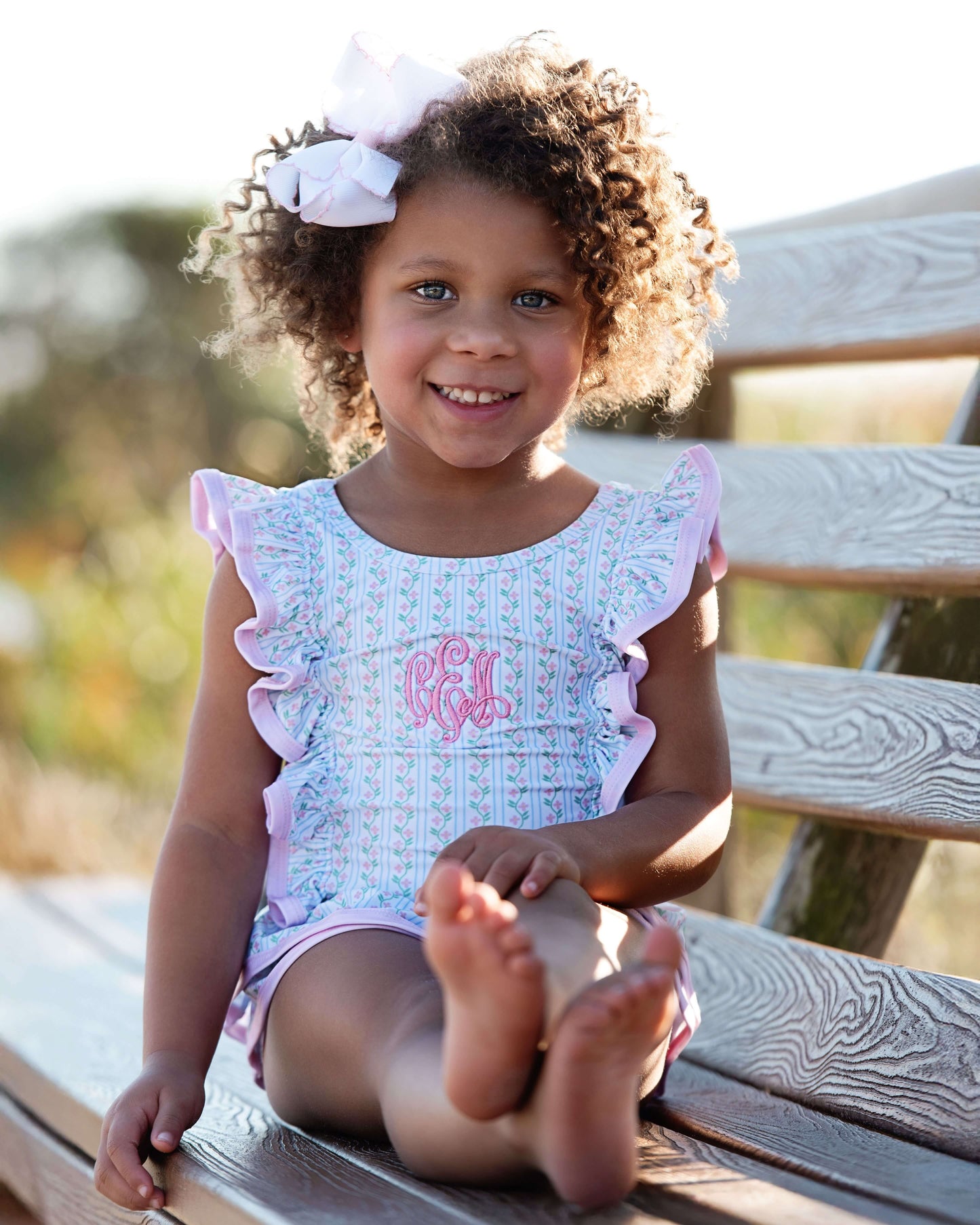 Young girl sitting on a wooden bench wearing a light blue dress with pink trim and a pink monogram.