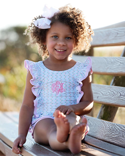 Young girl sitting on a wooden bench wearing a light blue dress with pink trim and a pink monogram.
