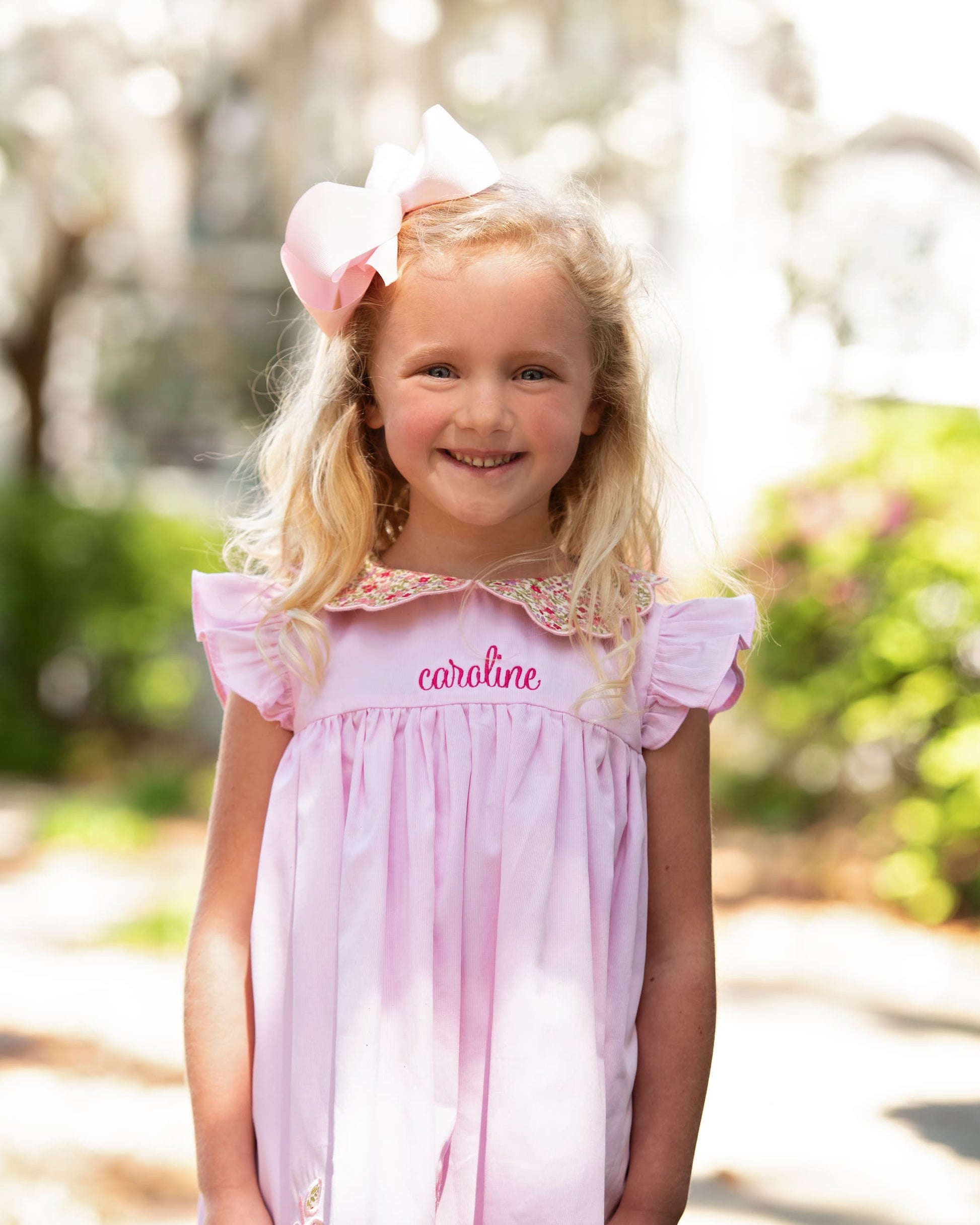 Young girl smiling wearing a pink bow and pink dress with the name caroline on it