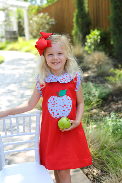 Young girl standing next to a chair holding an apple and wearing a red dress with floral big apple on the chest.