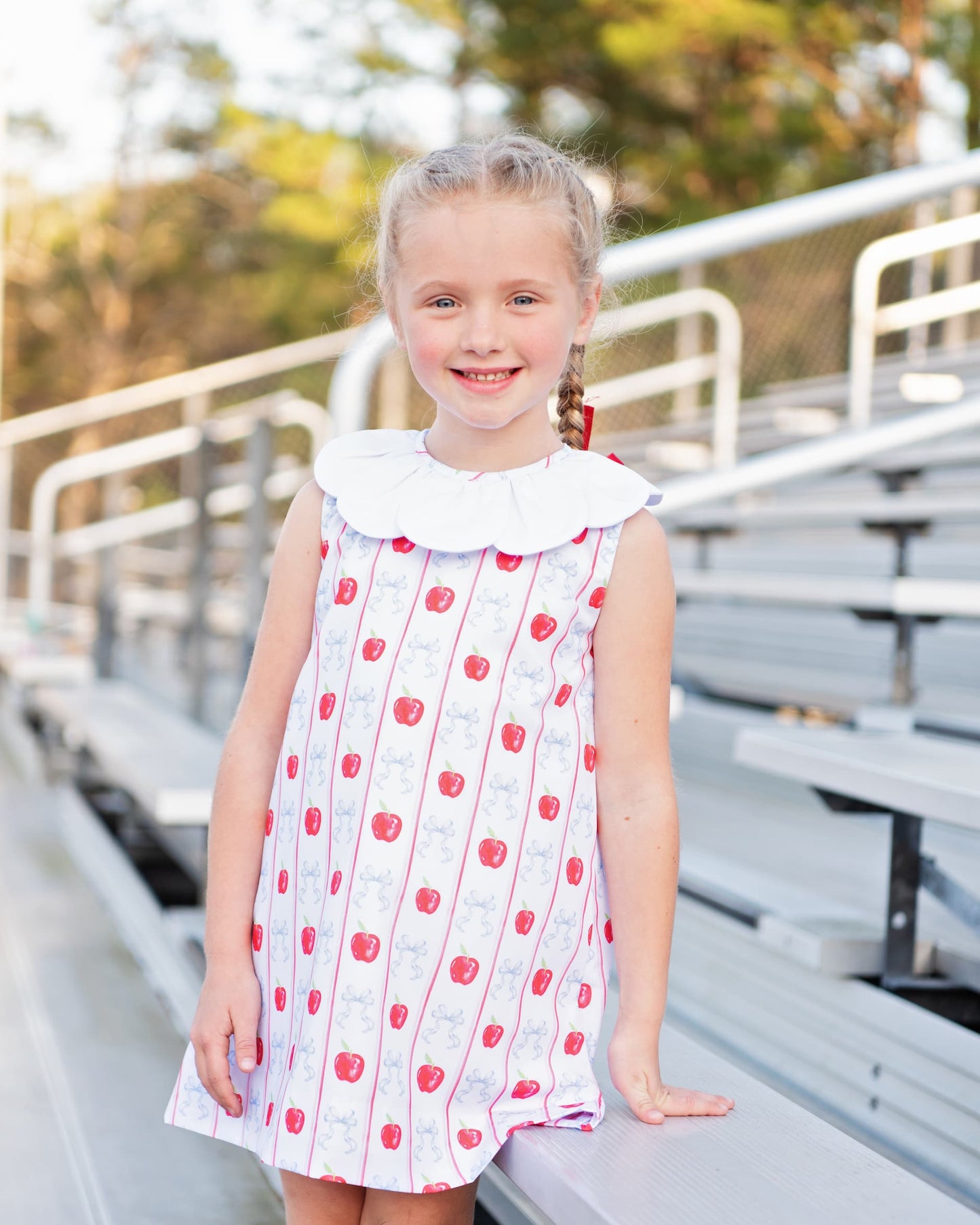 Young girl standing on bleachers wearing an apple and bow print dress.