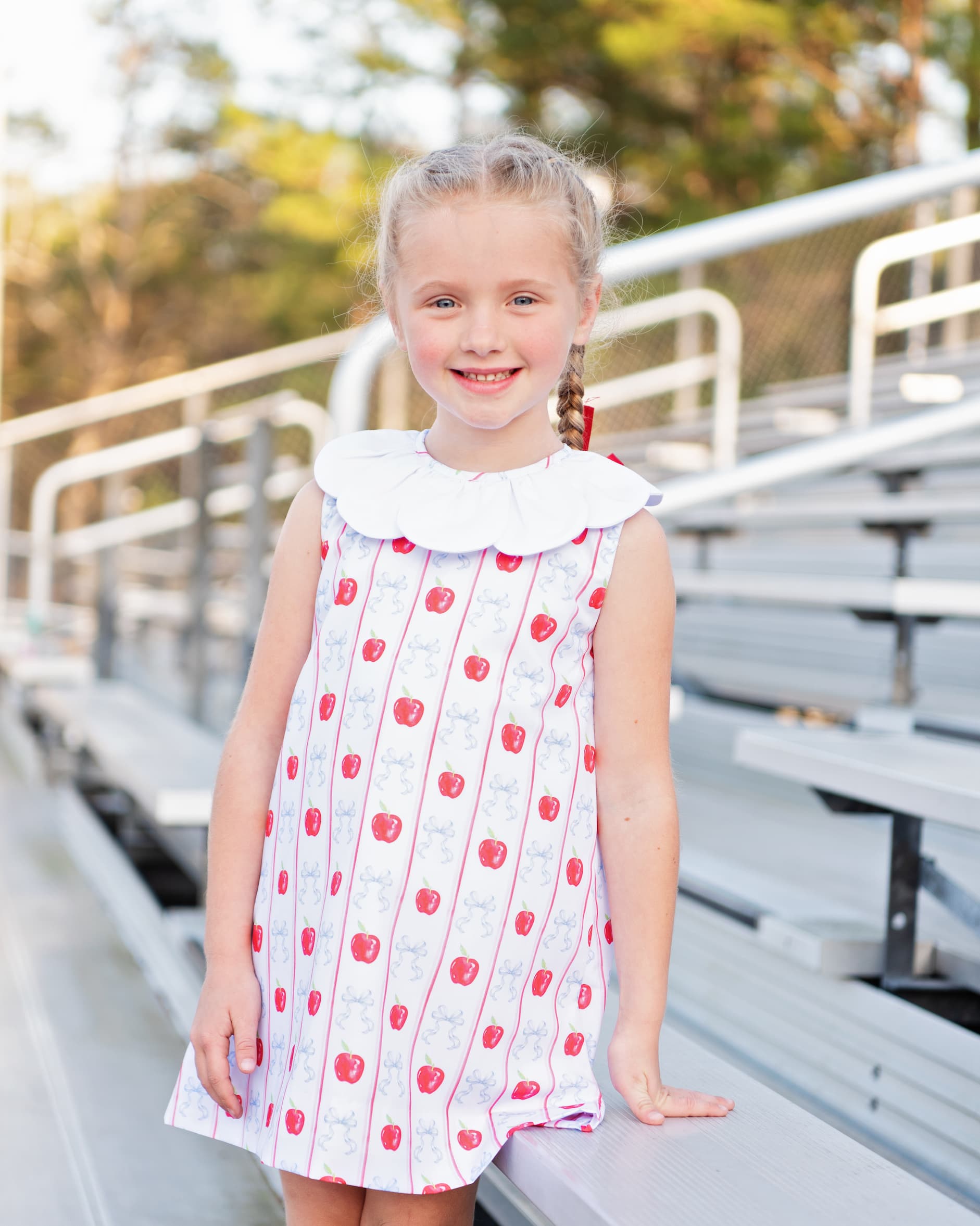 Young girl standing on bleachers wearing an apple and bow print dress.