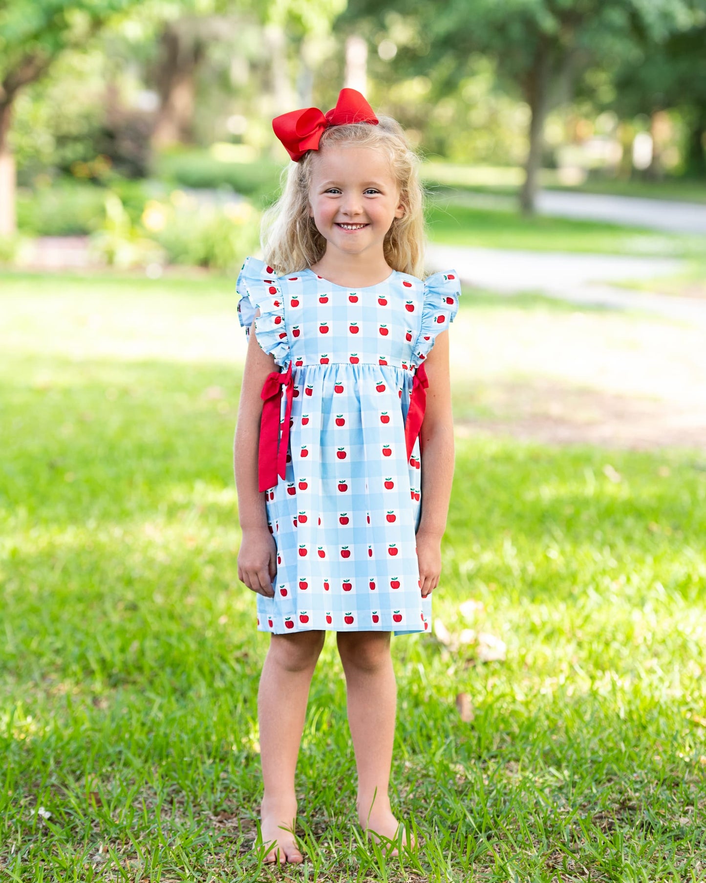 Young girl standing on grass wearing a blue dress with apples on it. 