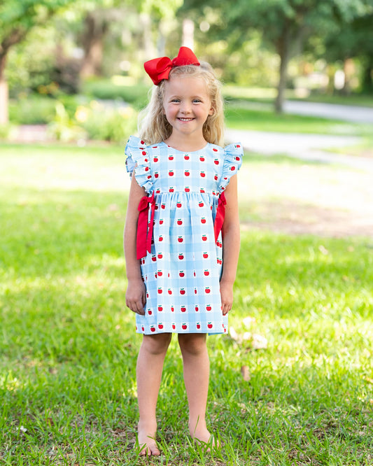 Young girl standing on grass wearing a blue dress with apples on it. 