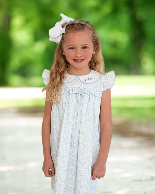 Young girl standing outside wearing a blue floral dress with a name monogrammed on the collar