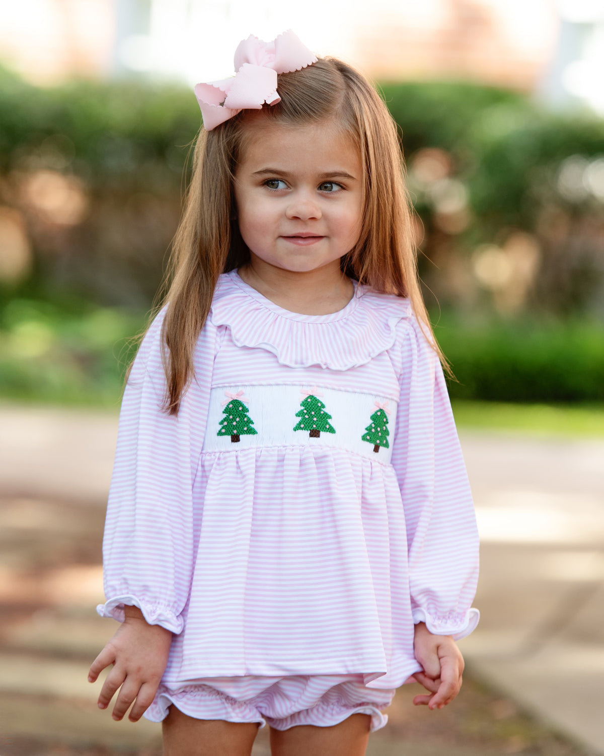 Young girl standing outside wearing a pink knit striped diaper set with smocked Christmas Trees and a matching pink bow
