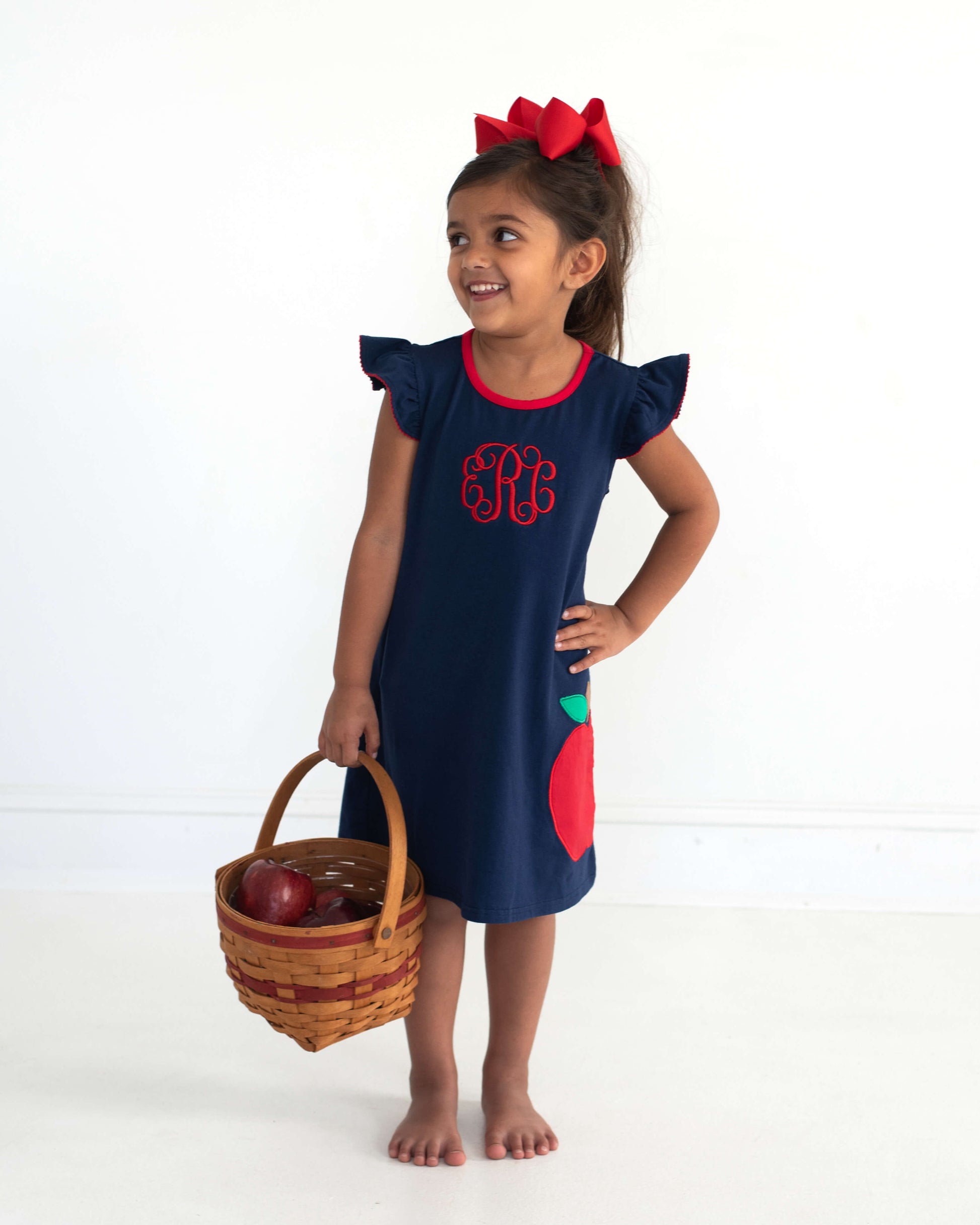 Young girl wearing a basket with apples in it wearing a navy dress with an applique apple.