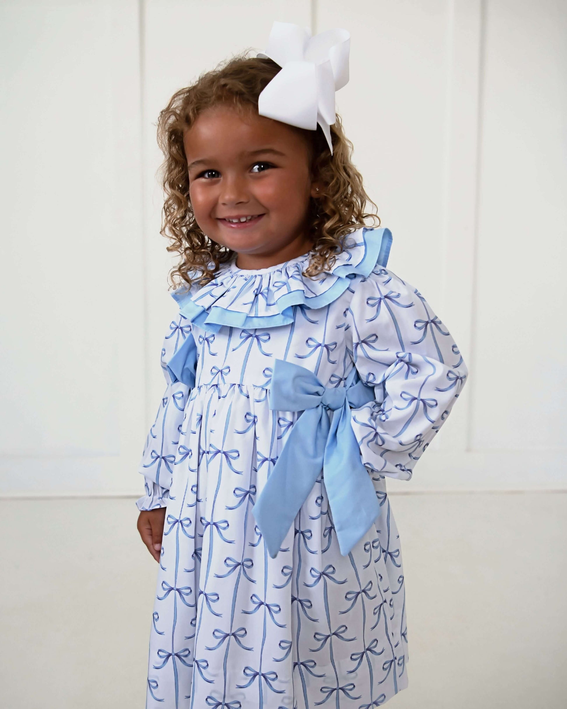 Young girl wearing a blue and white dress with a large bow, standing against a white background.