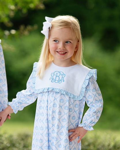 Young girl wearing a blue and white dress with a monogram, standing outdoors.