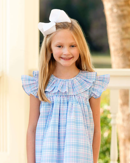 Young girl wearing a blue checkered dress with a white bow in her hair, standing outdoors.