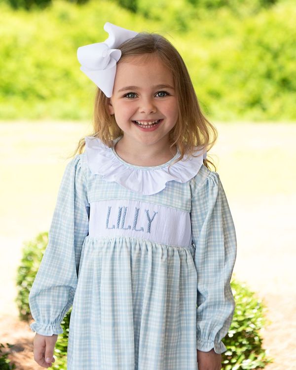 Young girl wearing a blue checkered dress with a white collar and a large white bow, standing outdoors.