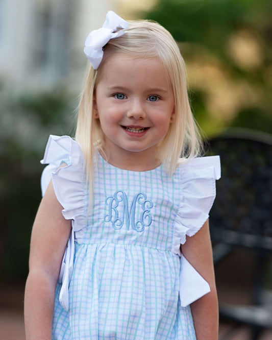 Young girl wearing a blue checkered dress with white ruffles and a large bow in her hair.