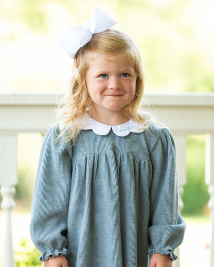 Young girl wearing a blue dress with a white collar and bow in her hair, standing outdoors.
