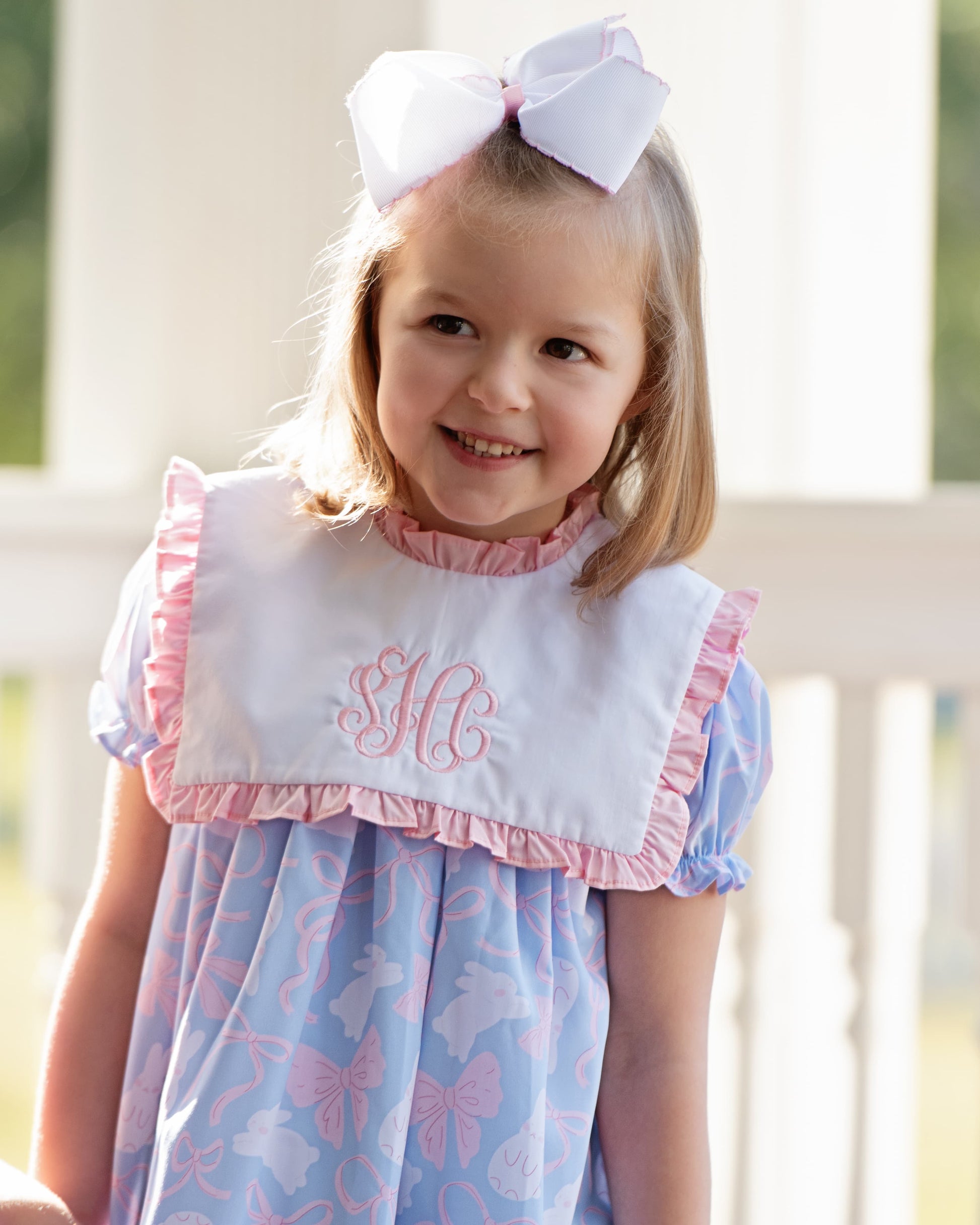 Young girl wearing a blue dress with pink trim and a white bow, standing outdoors.