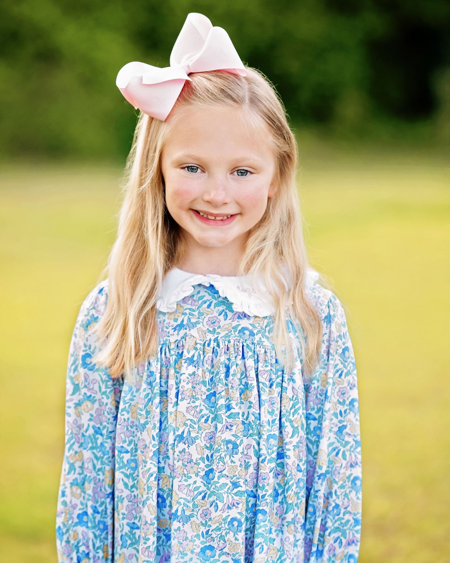 Young girl wearing a blue floral dress with a large pink bow in her hair, standing outdoors.