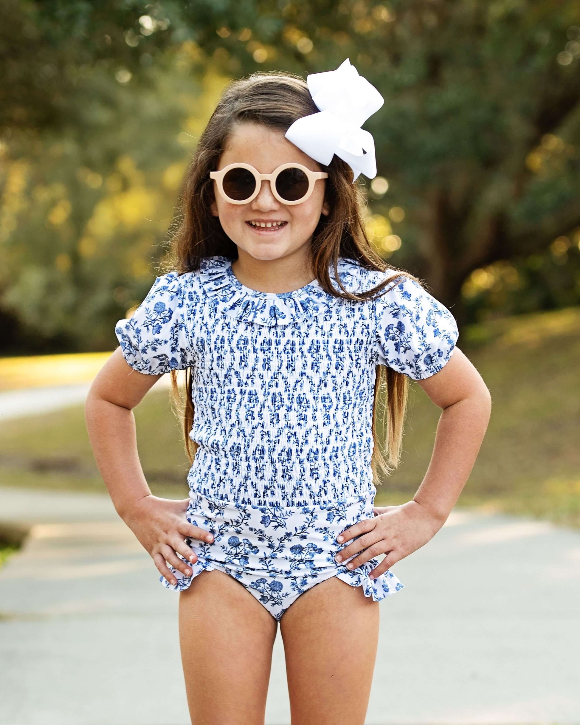 Young girl wearing a blue floral swimsuit with sunglasses and a white bow outdoors.