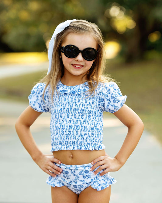 Young girl wearing a blue floral two-piece swimsuit and sunglasses outdoors.