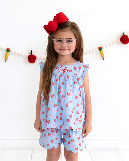 Young girl wearing a blue outfit with red apple pattern and a red bow, standing against a white background.