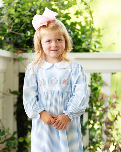Young girl wearing a blue striped dress with embroidered turkeys and a pink bow in her hair, standing outdoors.